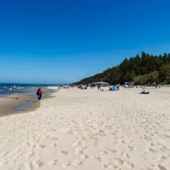 Słoneczna plaża w Sztutowie, z ludźmi spacerującymi brzegiem morza. W tle las i zabudowania, przypominające o bliskości aquaparku Sztutowo.