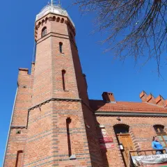 Ceglana latarnia morska w Ustce z charakterystyczną wieżą i budynkiem. Na balkonie stoi kobieta.
