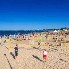 Plaża Brzeźno tętni życiem. Ludzie grają w siatkówkę plażową, opalają się i spacerują. W tle widać morze i namioty.