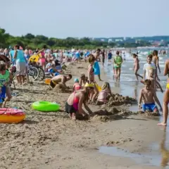 Tłoczna plaża jelitkowo w słoneczny dzień. Dzieci budują zamki z piasku, dorośli odpoczywają, a fale delikatnie obmywają brzeg.