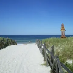 Piękna, piaszczysta lubiatowo plaża z drewnianym płotkiem i wydmami. W oddali widać morze i kilka osób kąpiących się.