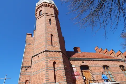 Ceglana latarnia morska w Ustce z charakterystyczną wieżą i budynkiem. Na balkonie stoi kobieta.