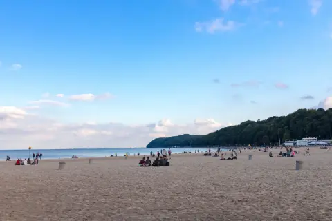 Ludzie odpoczywają na piaszczystej plaży miejskiej Gdynia, w tle las i błękitne niebo.