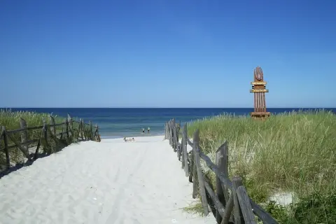 Piękna, piaszczysta lubiatowo plaża z drewnianym płotkiem i wydmami. W oddali widać morze i kilka osób kąpiących się.