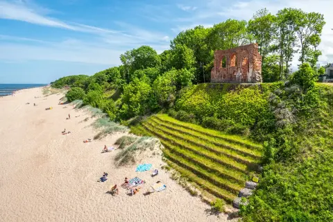 Słoneczna plaża w Trzęsaczu z ruinami kościoła na klifie. Piasek, morze i zielone wzgórze tworzą malowniczy krajobraz.