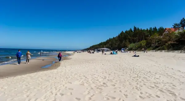 Słoneczna plaża w Sztutowie, z ludźmi spacerującymi brzegiem morza. W tle las i zabudowania, przypominające o bliskości aquaparku Sztutowo.