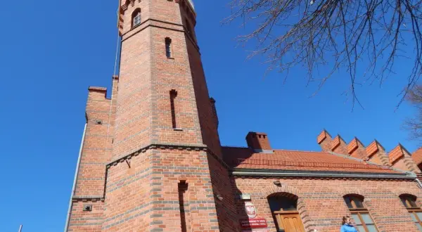 Ceglana latarnia morska w Ustce z charakterystyczną wieżą i budynkiem. Na balkonie stoi kobieta.
