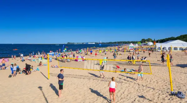 Plaża Brzeźno tętni życiem. Ludzie grają w siatkówkę plażową, opalają się i spacerują. W tle widać morze i namioty.