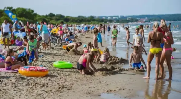 Tłoczna plaża jelitkowo w słoneczny dzień. Dzieci budują zamki z piasku, dorośli odpoczywają, a fale delikatnie obmywają brzeg.