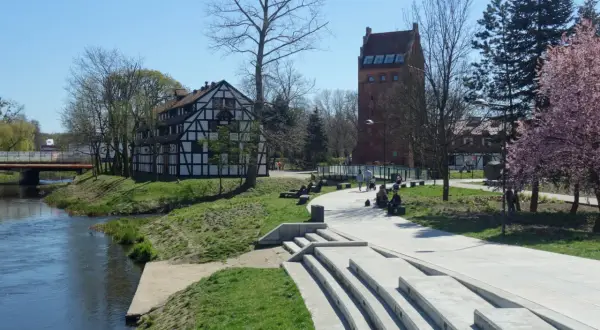 Słupsk atrakcje na weekend: nad rzeką widać zabytkową kamienicę i basztę, spacerują ludzie, kwitną drzewa.