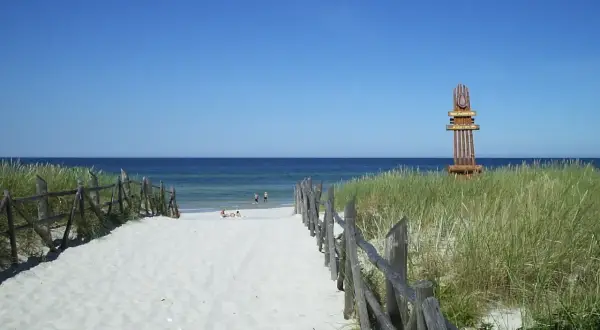 Piękna, piaszczysta lubiatowo plaża z drewnianym płotkiem i wydmami. W oddali widać morze i kilka osób kąpiących się.