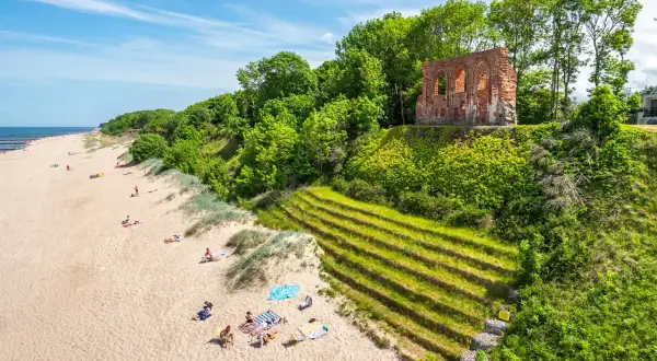 Słoneczna plaża w Trzęsaczu z ruinami kościoła na klifie. Piasek, morze i zielone wzgórze tworzą malowniczy krajobraz.