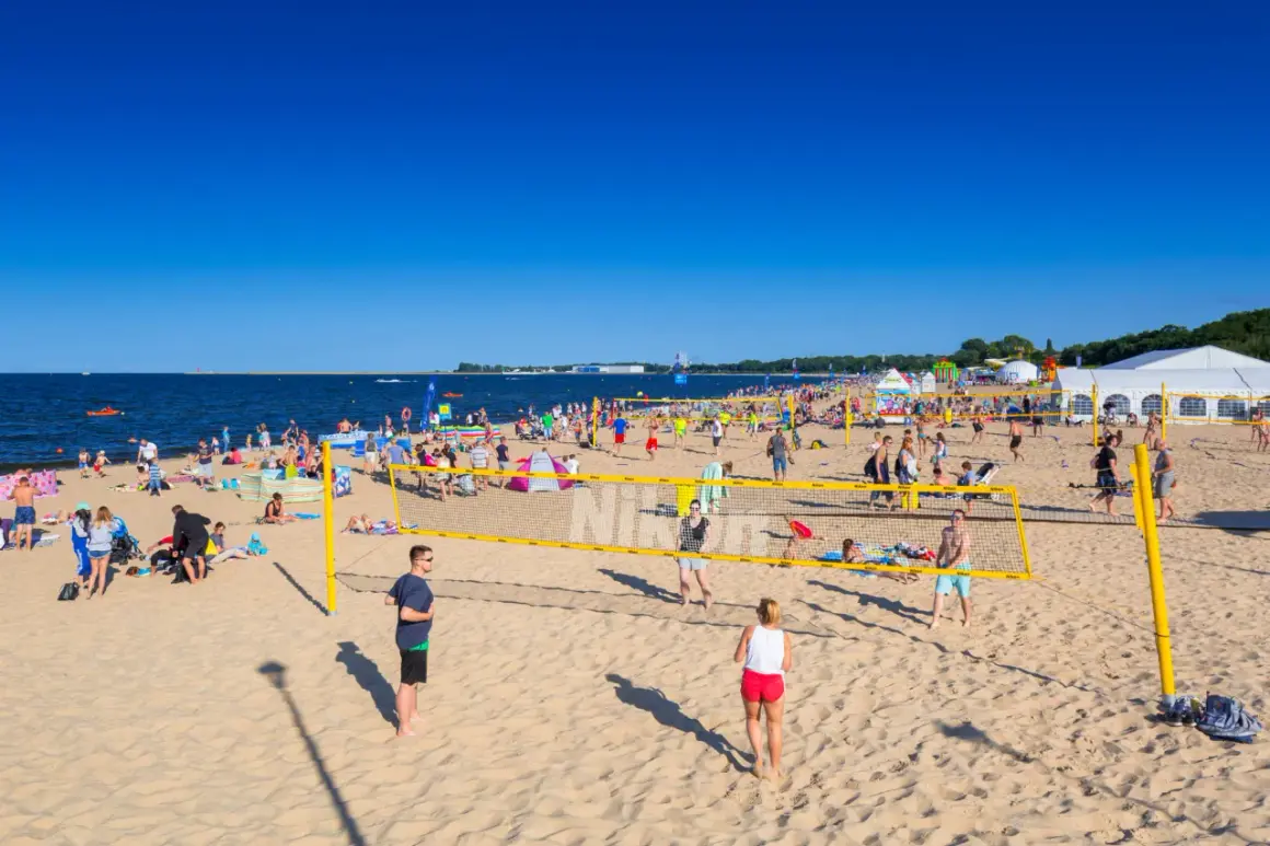 Plaża Brzeźno tętni życiem. Ludzie grają w siatkówkę plażową, opalają się i spacerują. W tle widać morze i namioty.