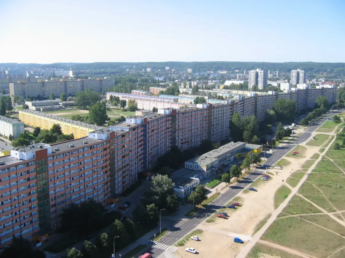 Długi, falisty budynek mieszkalny, znany jako falowiec gdańsk, ciągnie się przez krajobraz miejski, otoczony zielenią i innymi budynkami.