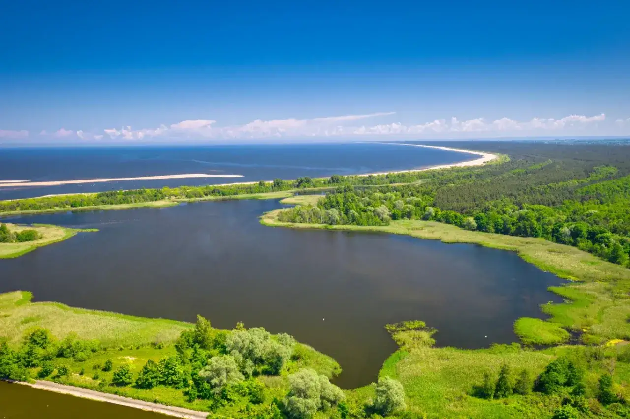 Malowniczy krajobraz z jeziorem, lasem i piaszczystą plażą. W oddali widać morze. To miejsce oferuje wiele atrakcji przyrodniczych.