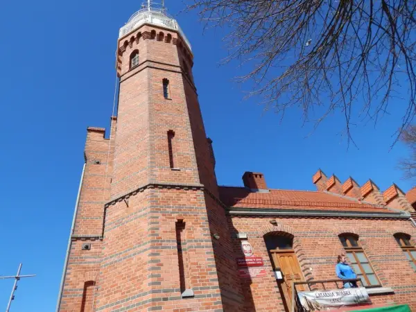 Ceglana latarnia morska w Ustce z charakterystyczną wieżą i budynkiem. Na balkonie stoi kobieta.
