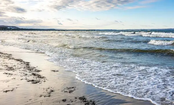 Plaża z falami i wodorostami, w oddali widać molo. To urokliwe kąpielisko w powiecie puckim zaprasza do spacerów.