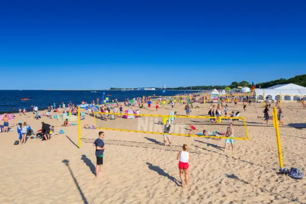 Plaża Brzeźno tętni życiem. Ludzie grają w siatkówkę plażową, opalają się i spacerują. W tle widać morze i namioty.