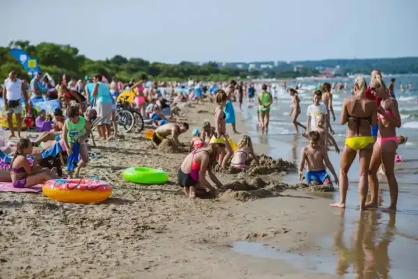 Tłoczna plaża jelitkowo w słoneczny dzień. Dzieci budują zamki z piasku, dorośli odpoczywają, a fale delikatnie obmywają brzeg.