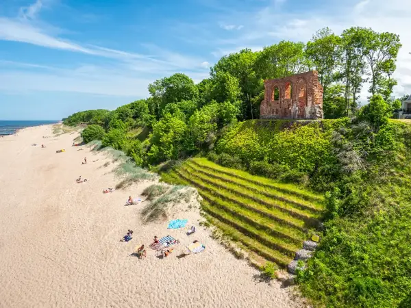 Słoneczna plaża w Trzęsaczu z ruinami kościoła na klifie. Piasek, morze i zielone wzgórze tworzą malowniczy krajobraz.